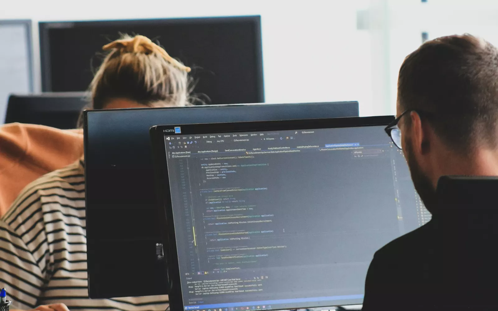 Two engineers at their desks in a bright open-plan office, one reviewing code on a large monitor